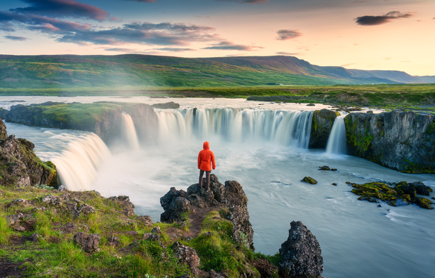 Godafoss waterfall flowing with colorful sunset sky and male tourist standing on cliff in summer at Iceland