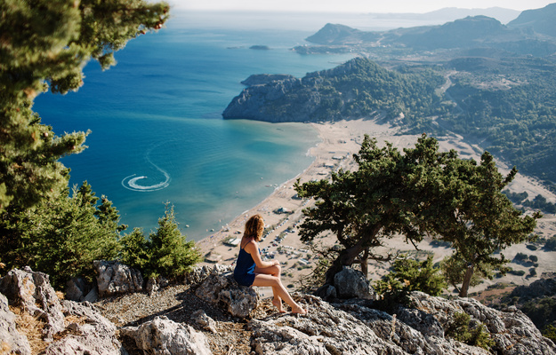 Photo of a sea landscape, view of Tsambika Bay on the island of Rhodes, Dodecanese, Greece. Girl sits on a stone and looks at a beautiful sandy beach with clear blue water. Jet ski draws patterns